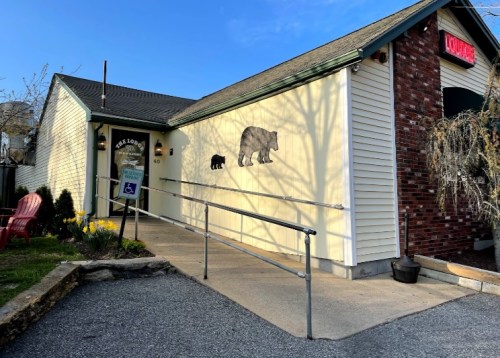 A building with a bear mural on the wall, featuring a ramp and red chairs outside, surrounded by greenery.