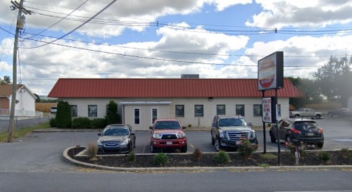 A one-story building with a red roof, surrounded by parked cars and shrubs, under a cloudy sky.