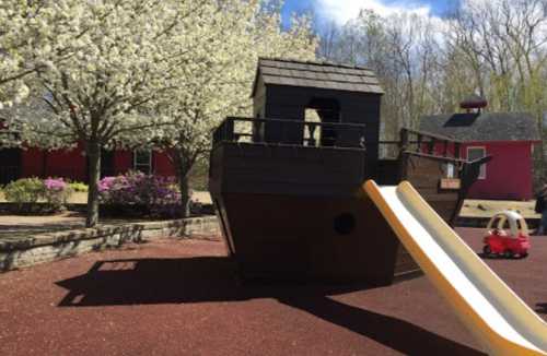 A playground featuring a wooden ship structure, a slide, and a small red toy car, surrounded by blooming trees.