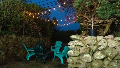 A cozy outdoor seating area with teal chairs and string lights, surrounded by stone walls and greenery at dusk.