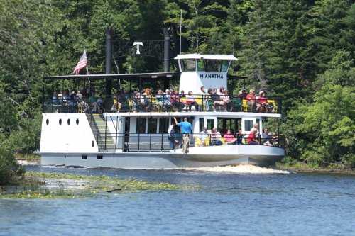 A large boat with passengers cruising on a calm river, surrounded by lush green trees. An American flag is visible.