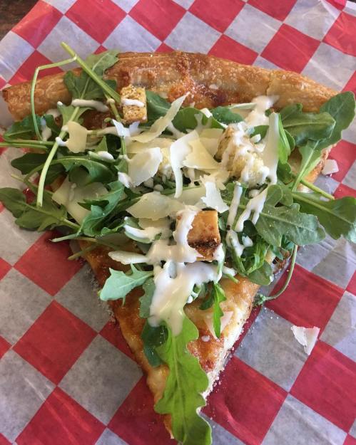 A slice of pizza topped with arugula, grilled chicken, and drizzled with creamy dressing on a red-checkered paper.