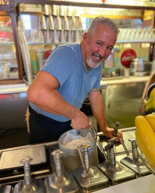 A smiling man scoops ice cream at a vintage ice cream shop, surrounded by glassware and colorful bananas.
