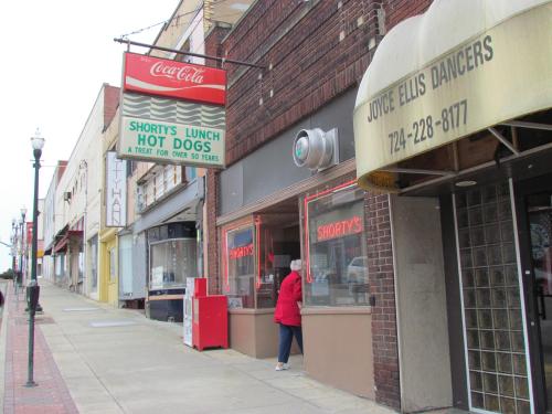 A street view featuring Shorty's Lunch hot dog stand, with a red sign and nearby shops on a cloudy day.
