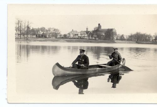 Two men in a canoe on a calm lake, surrounded by trees and distant buildings, captured in a vintage black-and-white photo.
