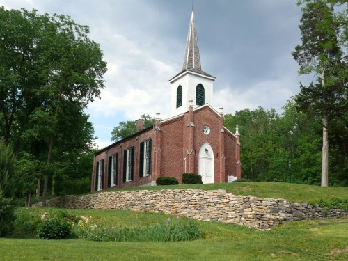 A brick church with a tall steeple, surrounded by greenery and a stone wall, under a cloudy sky.