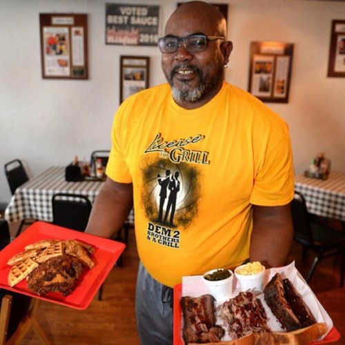 A man in a yellow "License to Grill" shirt holds two plates of barbecue food in a restaurant setting.