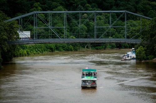 A boat navigates a river beneath a metal bridge, surrounded by lush greenery.
