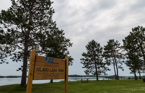 Sign for Island Lake Park near a calm lake, surrounded by tall pine trees under a cloudy sky.