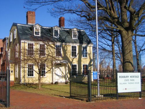 Historic yellow house with a green roof, surrounded by trees, at Roxbury Heritage State Park. Sign visible in front.