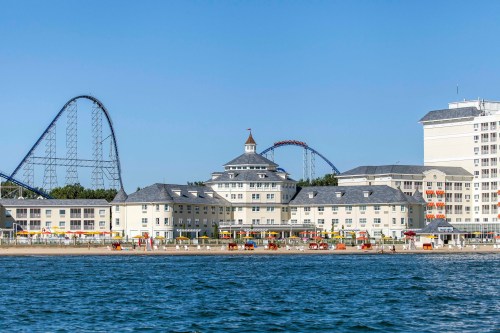 A seaside view featuring a hotel, beach, and roller coasters in the background under a clear blue sky.