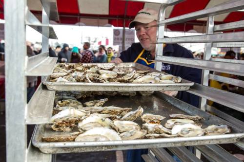 A vendor serves fresh oysters from trays at a bustling outdoor market under a red and white tent.
