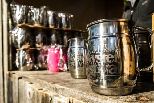 Metal mugs with engraved text sit on a wooden table, with more mugs stacked in the background.