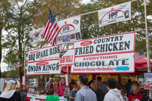 Food stalls at a fair featuring BBQ ribs, onion rings, fried chicken, and chocolate treats, with flags in the background.