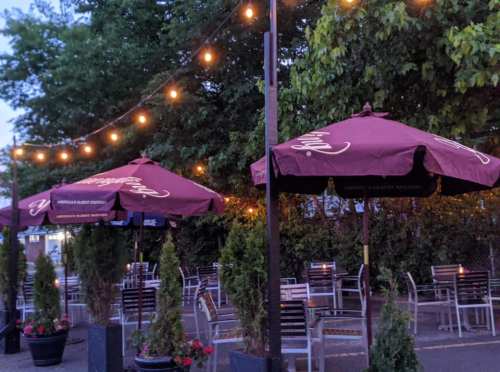 Outdoor seating area with maroon umbrellas, string lights, and greenery in the background during twilight.