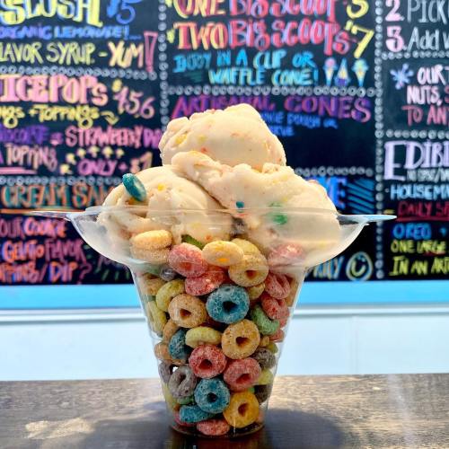 A clear cup filled with colorful cereal and topped with scoops of ice cream, set against a vibrant menu board.