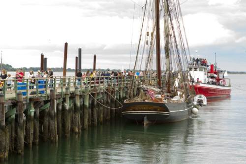 A historic ship docked at a pier with people walking along the waterfront under a cloudy sky.