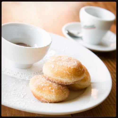 A plate with three sugar-dusted doughnuts and a small cup of dipping sauce, alongside a coffee cup.