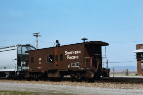 A Southern Pacific caboose on a railway track, with a freight car and a building in the background under a clear sky.