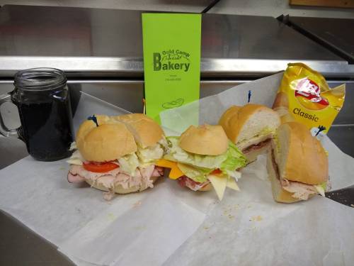 Three sandwiches with various fillings, a jar of drink, and a bag of chips on a countertop, with a bakery menu in the background.