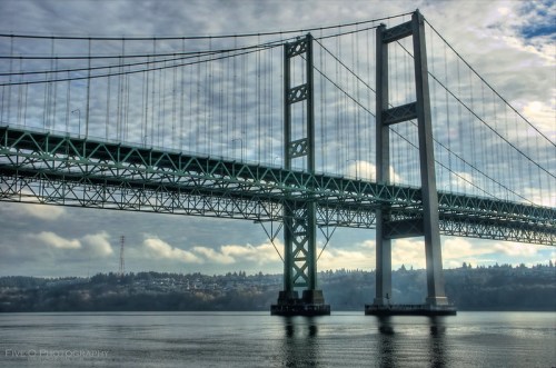 A large suspension bridge spans a calm body of water, with a cloudy sky and trees in the background.