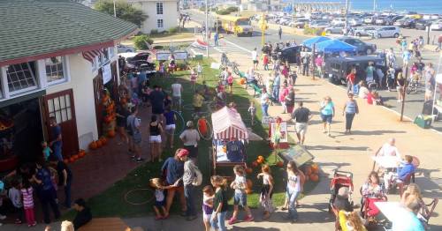 A bustling outdoor event with people enjoying activities, food stalls, and pumpkins near the beach on a sunny day.