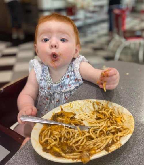 A messy baby with red hair enjoys spaghetti at a diner, with sauce on their face and food on the plate.