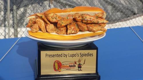A large sandwich filled with grilled chicken pieces, displayed on a trophy base labeled "Presented by Lupo's Spiedies."