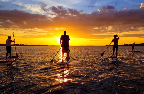 Four people paddleboarding on a calm lake at sunset, with vibrant orange and purple skies reflecting on the water.