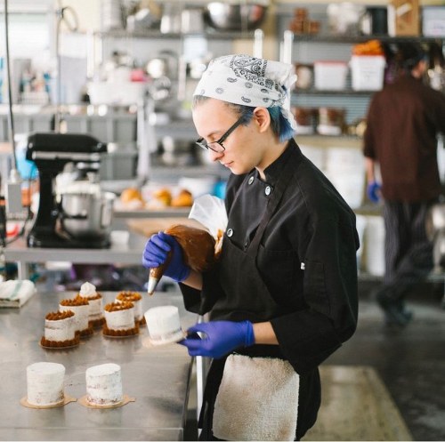 A baker in a black chef's coat decorates small cakes in a busy kitchen, wearing gloves and a bandana.