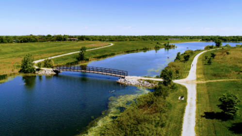 Aerial view of a serene landscape with a bridge over a lake, surrounded by green fields and walking paths.