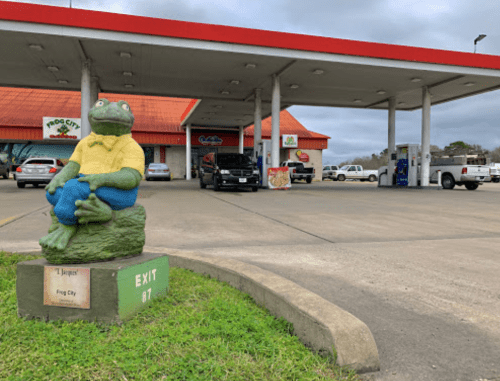 A large frog statue in a yellow shirt sits outside a gas station with a red roof and various vehicles parked nearby.