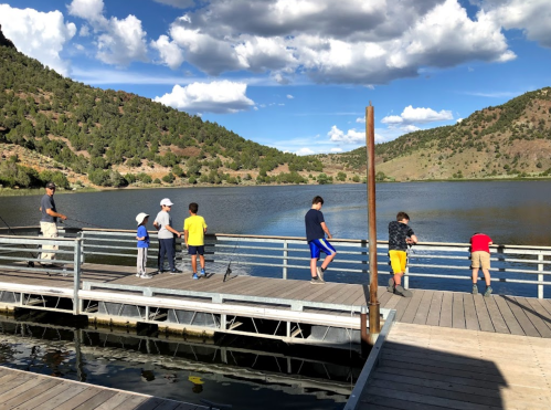 A group of children and an adult fish from a wooden dock by a lake, surrounded by hills and blue skies.