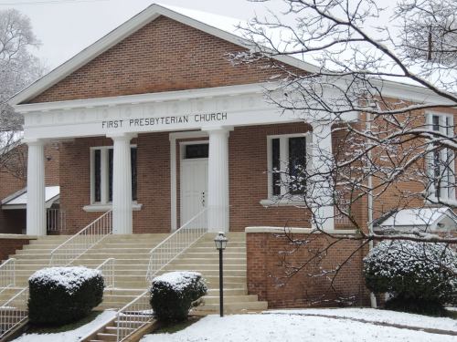 First Presbyterian Church building with snow covering the ground and branches, featuring white columns and brick facade.