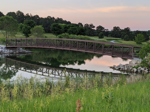 A wooden bridge arches over a calm pond, reflecting the surrounding greenery and a pastel sky at dusk.