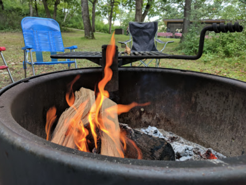 A fire pit with flames and logs, surrounded by camping chairs in a wooded area.