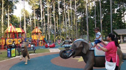 A child rides an elephant statue in a colorful playground surrounded by trees, with a lion statue nearby.