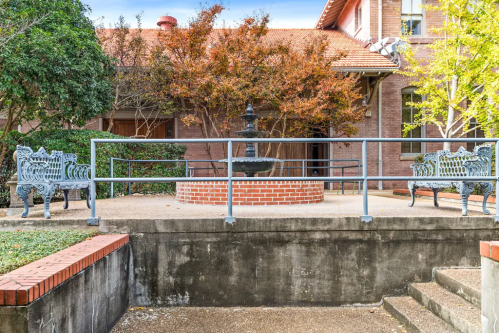 A courtyard with a fountain surrounded by benches and trees, featuring a brick wall and steps leading down.
