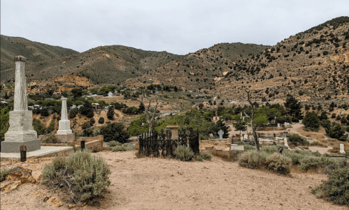 A cemetery with gravestones and a wrought iron fence, set against a backdrop of mountains and cloudy skies.