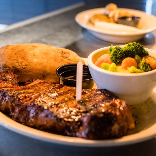 A plate featuring a grilled steak, baked potato, and a side of mixed vegetables in a bowl.
