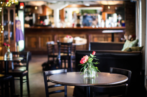 A cozy café interior with wooden tables, a vase of flowers, and a bar in the background. Soft lighting creates a warm atmosphere.
