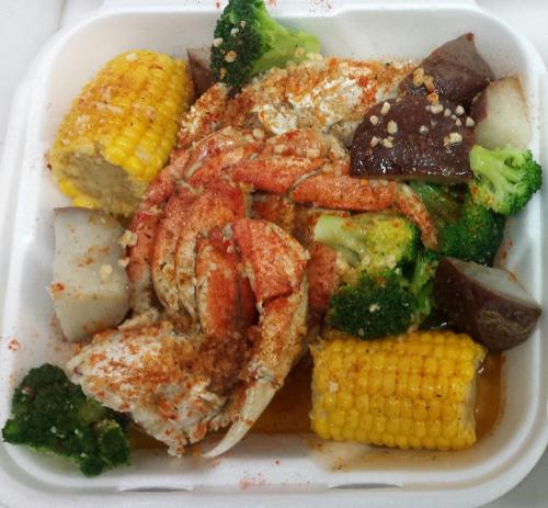 A takeout container filled with crab, corn, broccoli, and potatoes, topped with seasoning and spices.