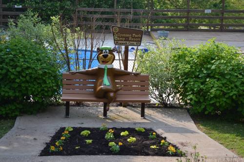 A bear statue in a tie sits on a bench near a flower bed and a swimming pool in a campground setting.