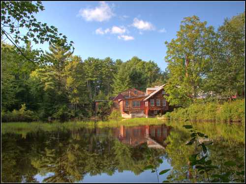 A serene pond reflects a rustic cabin surrounded by lush trees and a clear blue sky.