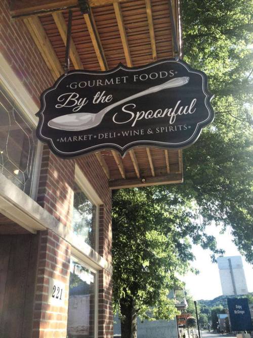 Sign for "By the Spoonful" featuring gourmet foods, market, deli, wine, and spirits, set against a brick building.