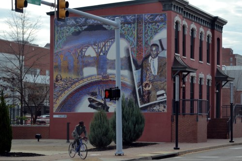 A cyclist passes by a colorful mural depicting a musician on a building, with traffic lights and trees nearby.