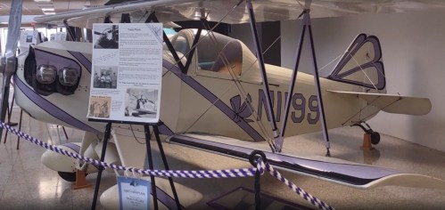 A vintage biplane displayed indoors, with a sign detailing its history and a purple and white color scheme.