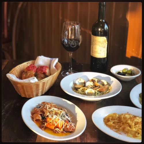 A table setting with pasta dishes, a basket of bread, olives, a glass of red wine, and a bottle of wine.
