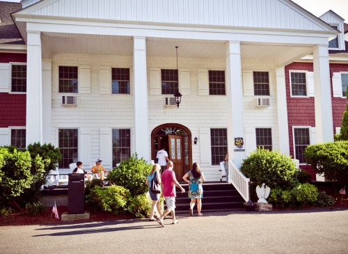 A group of people walks towards a large, red and white building with a porch and greenery around it.