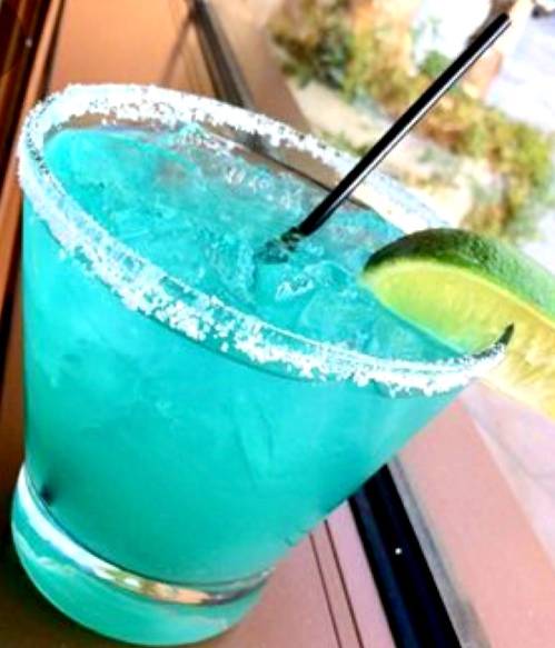 A vibrant blue cocktail in a glass with a salted rim and a lime wedge, served with a black straw.